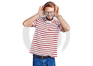 Handsome blond man with beard wearing casual clothes and glasses trying to hear both hands on ear gesture, curious for gossip