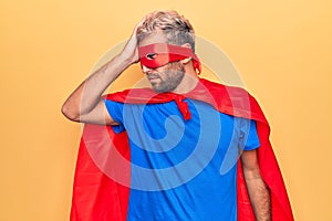 Handsome blond man wearing super hero costume with mask and cape over yellow background surprised with hand on head for mistake,