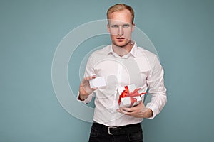 Handsome blonde man wearing white shirt isolated over blue background wall holding credit card and gift box looking at