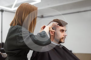 Handsome blue eyed man sitting in barber shop. Hairstylist Hairdresser Woman cutting his hair. Female barber