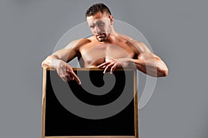 Handsome bodybuilder man with sign advertising board. Strong muscular athlete holding blank black poster ad. Copyspace.