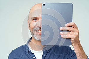 Handsome bold man cover half of his face with digital tablet in hand smiling looking at camera isolated on white