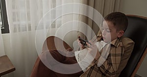 Handsome boy of 6 years old plays a mobile game on a smartphone at home while sitting in an armchair. Adorable kid boy