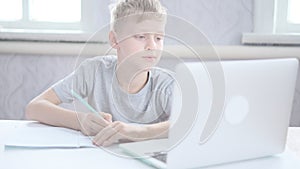handsome boy does his homework and writes in notebook sitting at table in front of laptop. Online training. Remote