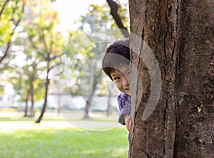 Handsome boy hiding behind tree for playing hide and seek with friend at park or son hiding mother behind tree because little kid