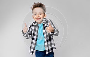 A handsome boy in a plaid shirt, blue shirt and jeans stands on a gray background. The boy smiles and shows class