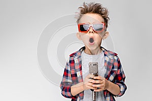 A handsome boy in a plaid shirt, gray shirt and jeans stands on a gray background. A boy wearing sunglasses. Red-haired