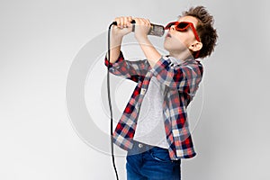 A handsome boy in a plaid shirt, gray shirt and jeans stands on a gray background. A boy wearing sunglasses. Red-haired