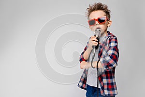 A handsome boy in a plaid shirt, gray shirt and jeans stands on a gray background. A boy wearing sunglasses. Red-haired