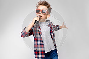 A handsome boy in a plaid shirt, gray shirt and jeans stands on a gray background. A boy wearing sunglasses. Red-haired