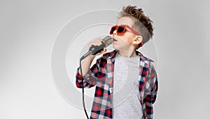 A handsome boy in a plaid shirt, gray shirt and jeans stands on a gray background. A boy wearing sunglasses. Red-haired