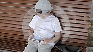 Handsome boy playing online games on smartphone outdoors. Kid boy wears cap and white t-shirt. Child sits on bench, looking some v