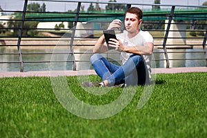 Handsome boy using tablet and headphones, drinking coffee to go, man sitting on grass and enjoying nice sunny day by river side.