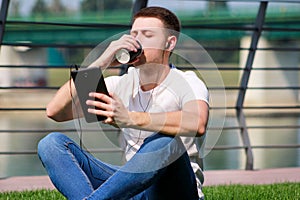 Handsome boy using tablet and headphones, drinking coffee to go, man sitting on grass and enjoying nice sunny day by river side.