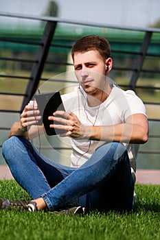 Handsome boy using tablet and headphones, drinking coffee to go, man sitting on grass and enjoying nice sunny day by river side.