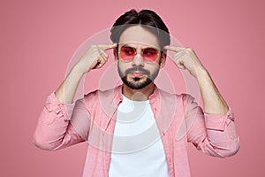 Handsome boy wearing stylish outfit and sunglasses looking away with thoughtful expression, keeping index fingers on his head.