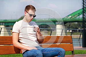 Handsome boy wearing sunglasses, sitting on a bench on a pier and drinking coffee to go. Taking a break from city rush.