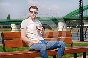 Handsome boy wearing sunglasses, sitting on a bench on a pier and drinking coffee to go. Taking a break from city rush.