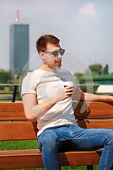 Handsome boy wearing sunglasses, sitting on a bench on a pier and drinking coffee to go. Taking a break from city rush.