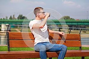 Handsome boy wearing sunglasses, sitting on a bench on a pier and drinking coffee to go. Taking a break from city rush.