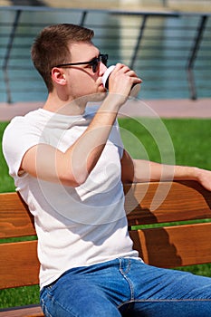 Handsome boy wearing sunglasses, sitting on a bench on a pier and drinking coffee to go. Taking a break from city rush.