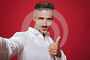 Handsome brunette man 30s with stubble in white shirt taking selfie photo and showing thumb up, isolated over red background