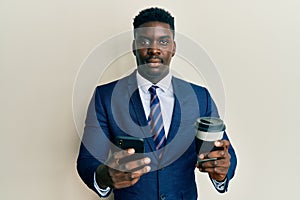 Handsome business black man using smartphone and drinking a cup of coffee relaxed with serious expression on face