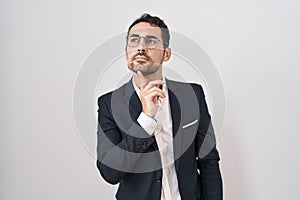 Handsome business hispanic man standing over white background thinking concentrated about doubt with finger on chin and looking up