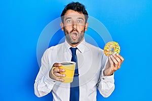 Handsome business man with beard eating doughnut and drinking coffee afraid and shocked with surprise and amazed expression, fear