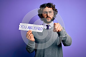 Handsome business man with beard holding you are fired message over purple background pointing with finger to the camera and to