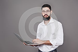 Handsome business man in glasses and suit holding laptop in hands and writing something. Side view. Isolated gray