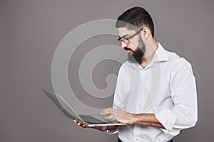 Handsome business man in glasses and suit holding laptop in hands and writing something. Side view. Isolated gray