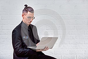 Handsome business man in glasses and suit holding laptop in hands and writing something. Side view. On white brick