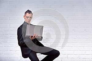 Handsome business man in glasses and suit holding laptop in hands and writing something. Side view. On white brick