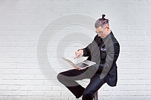 Handsome business man in glasses and suit holding laptop in hands and writing something. Side view. On white brick