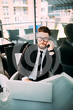 Handsome business man in suit relaxing outside with laptop at a terrace somewhere at a coffee shop talking at phone