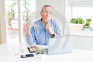 Handsome business senior man eating delivery sushi while working using laptop serious face thinking about question, very confused