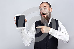 Handsome businessman with beard and handlebar mustache holding digital tablet and looking at camera and showing screen with finger