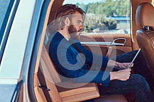 A handsome businessman with a beard and long hair sitting in the back seat of a luxury car and working with a tablet