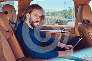 A handsome businessman with a beard and long hair sitting in the back seat of a luxury car and working with a tablet