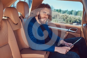 A handsome businessman with a beard and long hair sitting in the back seat of a luxury car and working with a tablet