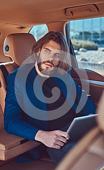 A handsome businessman with a beard and long hair sitting in the back seat of a luxury car and working with a tablet