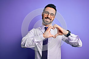 Handsome businessman with beard wearing casual tie and glasses over purple background smiling in love showing heart symbol and
