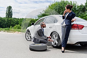 handsome businessman changing tires on car on road businesswoman talking