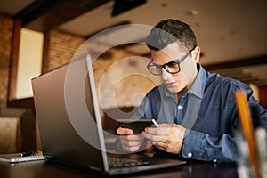 Handsome businessman distracted from work on the laptop watching video on smartphone. Freelancer holding mobile phone photo