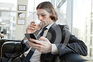 Handsome businessman drinking whiskey in office and checking text messages
