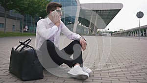 Handsome businessman in glasses at the airport sitting on the ground with luggage and talks by phone trying to solve the