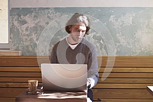 Handsome businessman hipster with long hair working on laptop in sunny cafe, having coffee in glass. Horizontal