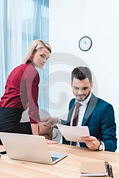 handsome businessman holding papers and touching knee of young female colleague sitting