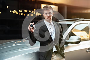 Handsome businessman leaning on his parked car with keys in his hand, young and successful